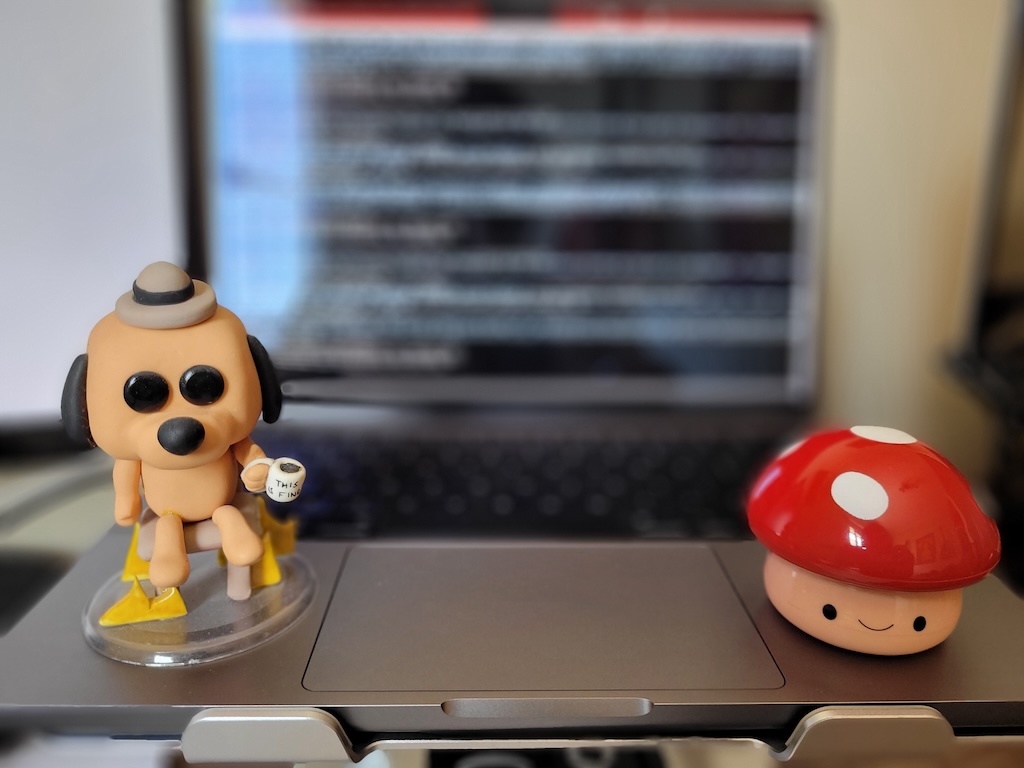 This Is Fine Dog Funko Pop and Mario's Super Mushroom sitting on a MacBook. This picture has a portrait effect, focused on the characters. The screen is blurry.